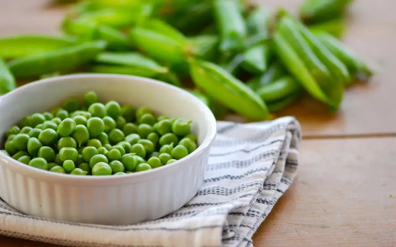 Una ciotola bianca piena di piselli freschi, un legume salutare, con baccelli sfocati sullo sfondo.