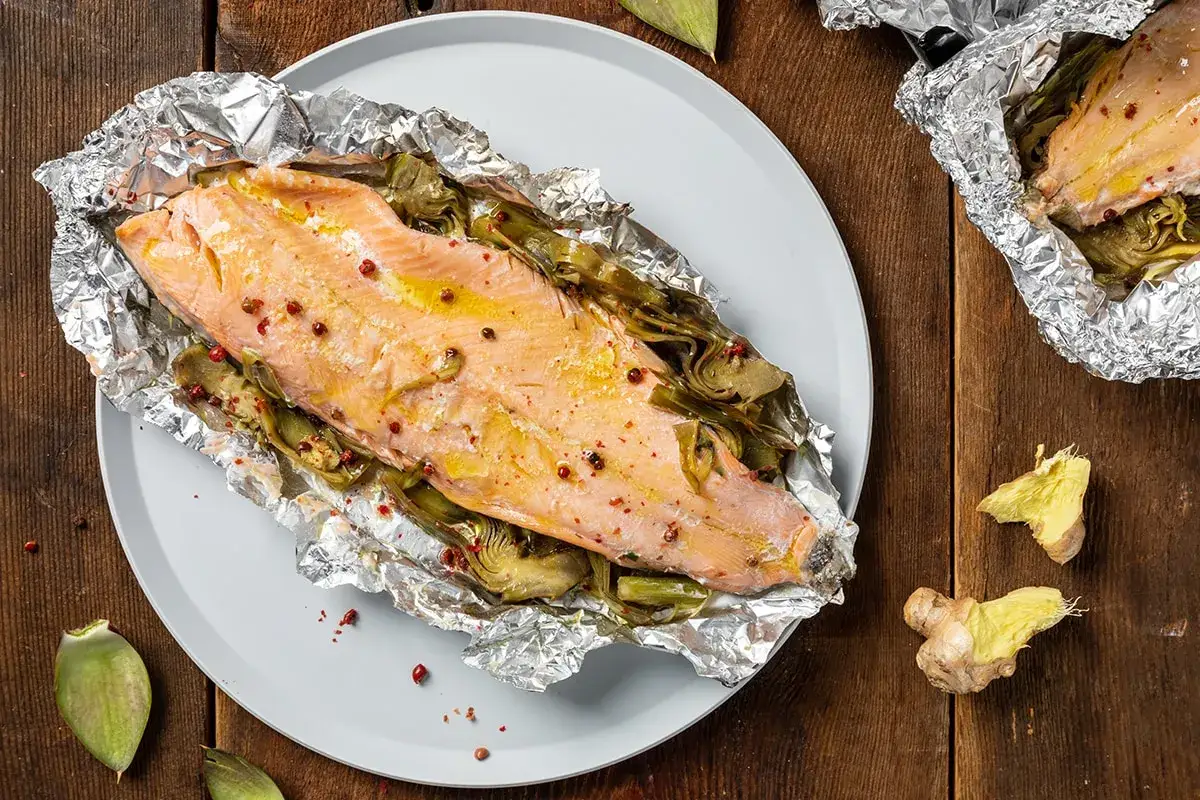 Trota salmonata cotta al cartoccio con carciofi e pepe rosa, servita su un piatto grigio su un tavolo di legno scuro.