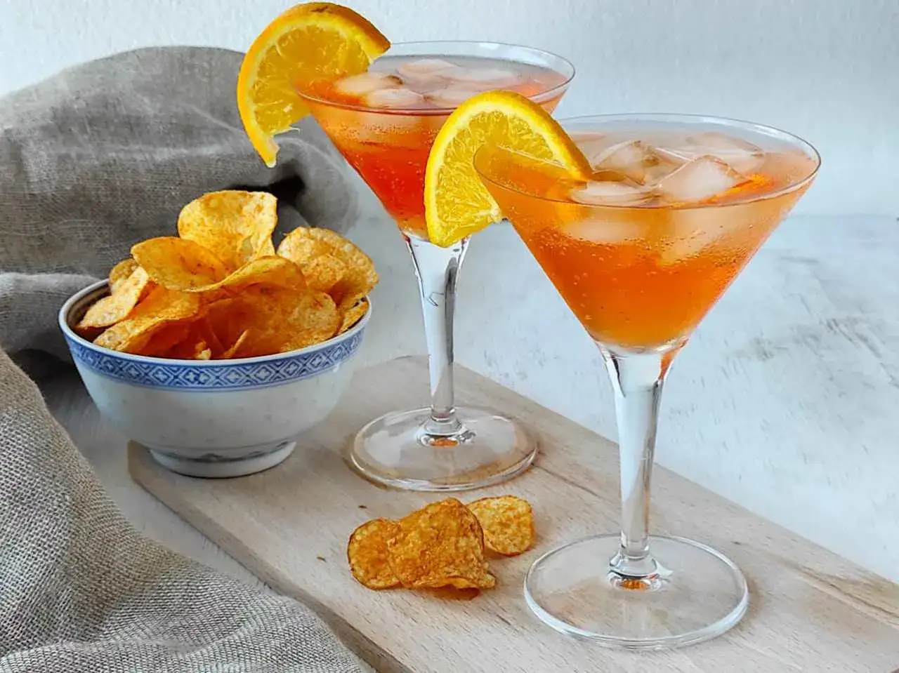 Due spritz arancioni con fette d'arancia e ghiaccio, accompagnati da una ciotola di patatine croccanti. Perfetti per un aperitivo, ma attenzione alle calorie spritz!