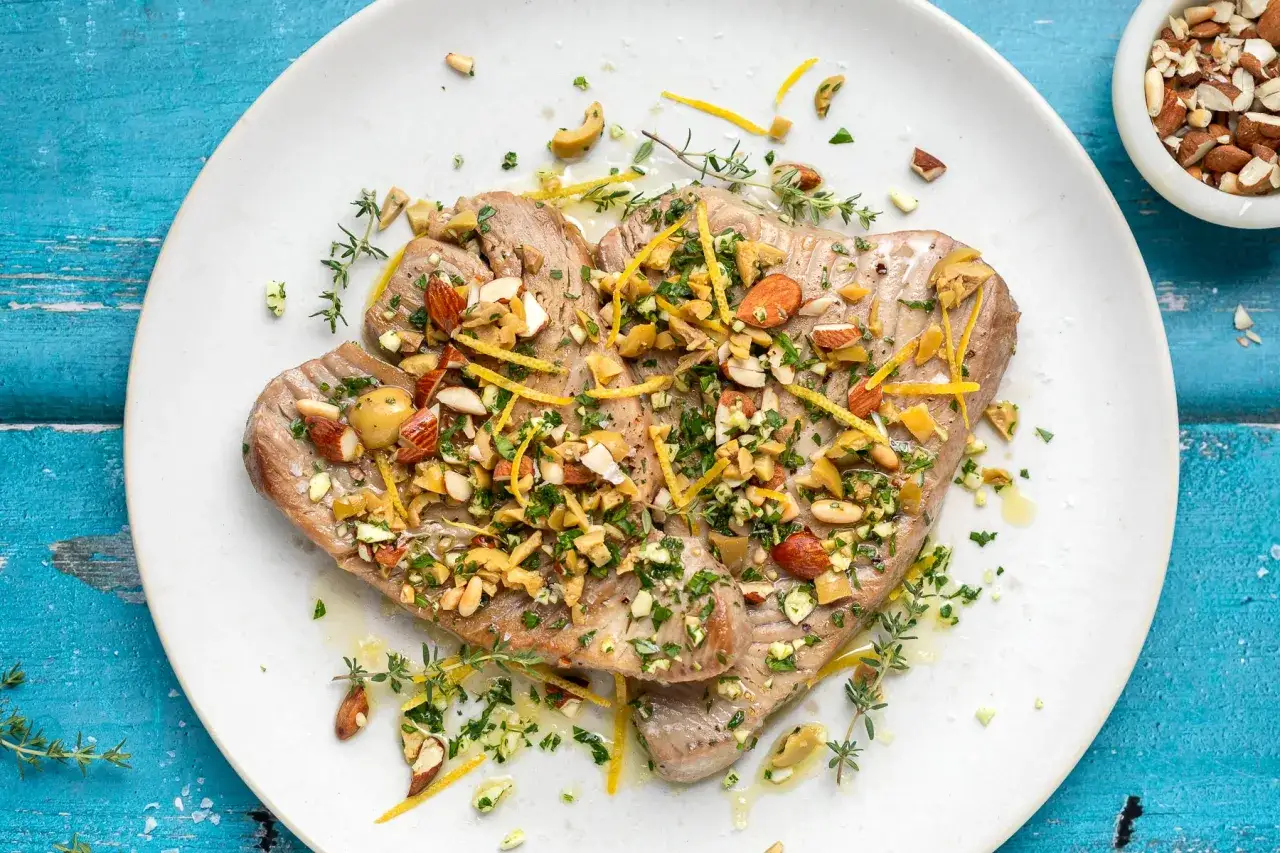 Delizioso tonno pesce cotto, guarnito con mandorle tritate, olive, scorza di limone e erbe aromatiche fresche su un piatto bianco.