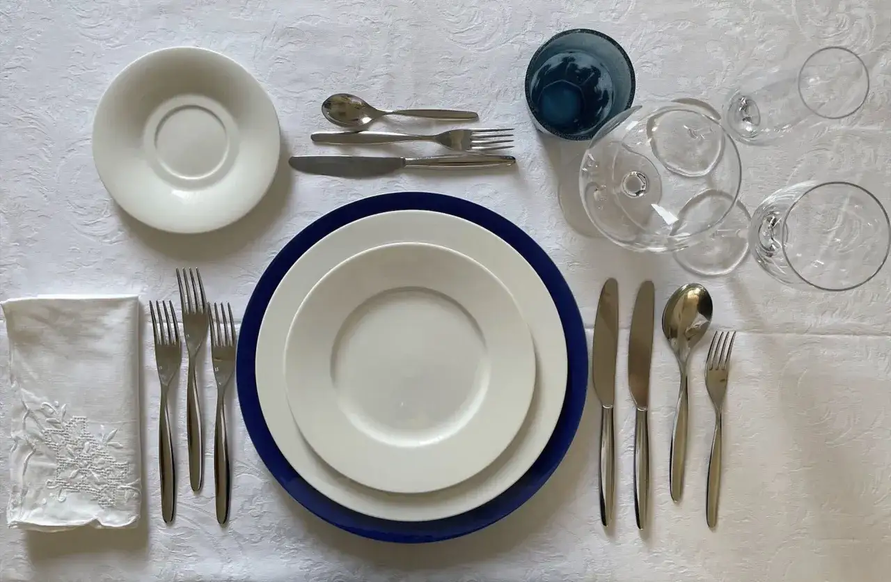 Tavola apparecchiata con piatti, posate e bicchieri. Si attende di sapere a chi si versa prima il vino.