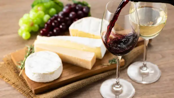 Un bicchiere di vino rosso viene versato, accanto a un bicchiere di vino bianco, con un assortimento di formaggi e uva su un tagliere.