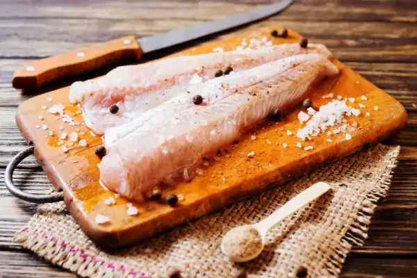 Filetti di nasello pesce pronti per essere cucinati, conditi con sale grosso e pepe su un tagliere di legno.
