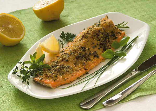 Filetto di trota salmonata cotto al forno con erbe aromatiche e limone, servito su un piatto bianco con guarnizioni fresche.