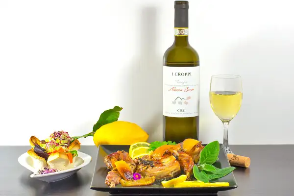 Piatto di coniglio arrosto con limone e erbe aromatiche, accompagnato da un bicchiere e una bottiglia di vino Albana Secco, perfetto vino da abbinare al coniglio.
