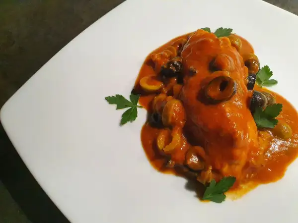 Un saporito pesce scorfano in salsa rossa con olive nere e verdi, guarnito con prezzemolo fresco su un piatto bianco.