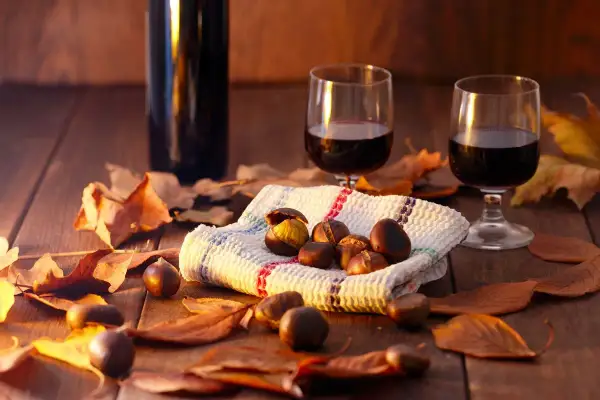 Vino per castagne su un tavolo di legno, circondato da foglie autunnali. Due bicchieri di vino rosso e una ciotola di castagne.