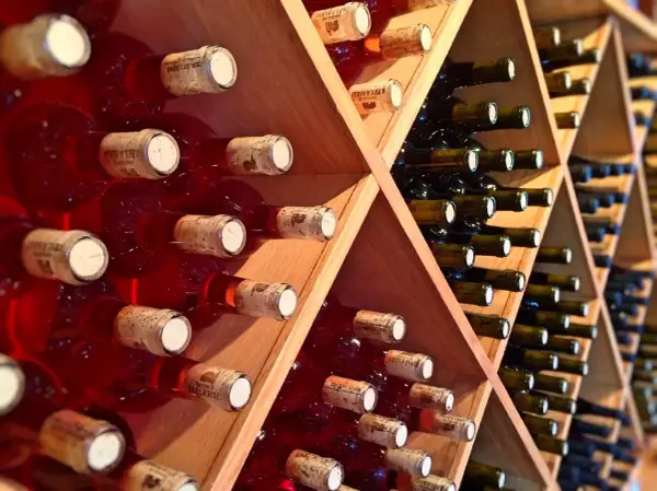 Scaffali di legno pieni di bottiglie di vino rosso, alcune con etichette visibili. Una piccola fioretta di vino attende di essere gustata.
