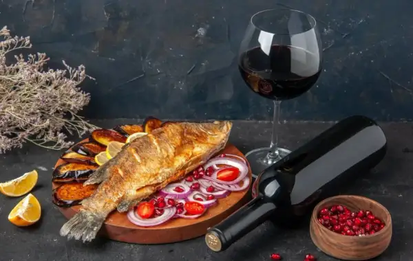 Un gustoso pesce fritto con melanzane, limone e cipolla, accompagnato da un bicchiere e una bottiglia di vino rosso da abbinare al pesce.