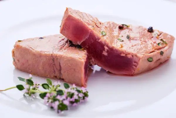 Due tranci di tonno pesce, conditi con erbe e pepe, su un piatto bianco, con un rametto di fiori viola in primo piano.