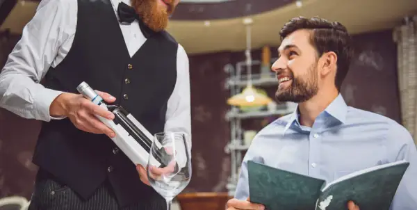 Cameriere con barba presenta una bottiglia di vino a un cliente sorridente che legge il menu. Si chiede a chi si versa prima il vino.