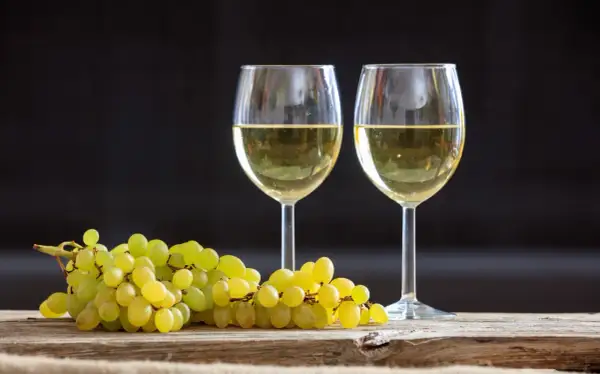 Due calici di vino bianco, tra i migliori vini bianchi italiani, accanto a un grappolo d'uva su un tavolo di legno.