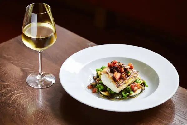 Un bicchiere di vino per pesce, accanto a un piatto di pesce bianco con verdure e pomodoro.